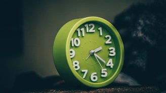 A green alarm clock with white numbers and hands stands against a dark, blurred background. The clock face displays the time as five minutes past ten. The image has a slightly dim and moody lighting, emphasizing the clock as the central focus.