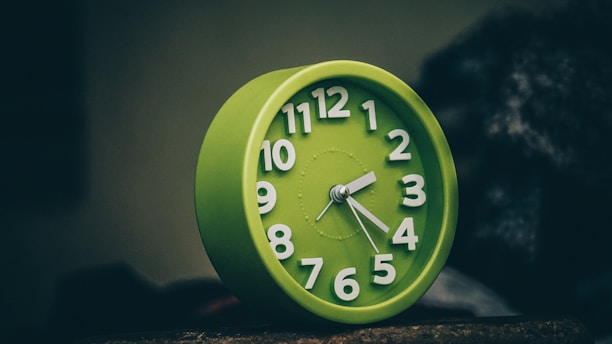 A green alarm clock with white numbers and hands stands against a dark, blurred background. The clock face displays the time as five minutes past ten. The image has a slightly dim and moody lighting, emphasizing the clock as the central focus.