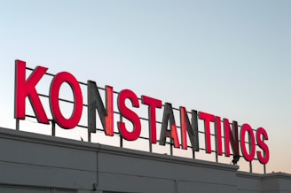 Large red neon letters spell out the word 'KONSTANTINOS' against a clear sky. The sign is mounted on a building rooftop.