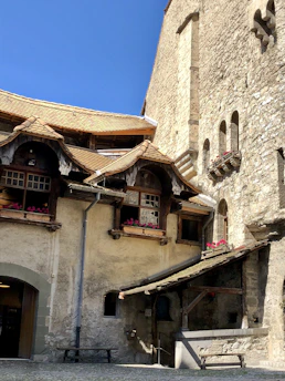 A charming, rustic building with stone walls and a shingled roof. The structure features wooden elements, small windows with window boxes filled with red flowers, and a small annex with an arched entrance. The stonework and wooden details suggest a historical or medieval architectural style.