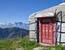 A colorful yurt with a detailed red door, set against a stunning backdrop of the snowy mountains and grassy hills on a clear day.
