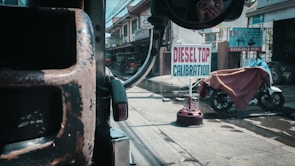 A street scene viewed from inside a vehicle, featuring a road sign for 'Diesel Top Calibration' prominently in the center. The foreground includes part of the vehicle's interior and side mirror. The surroundings show urban structures, a parked motorcycle covered with a tarp, and various commercial banners.