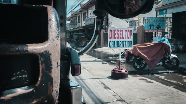 A street scene viewed from inside a vehicle, featuring a road sign for 'Diesel Top Calibration' prominently in the center. The foreground includes part of the vehicle's interior and side mirror. The surroundings show urban structures, a parked motorcycle covered with a tarp, and various commercial banners.