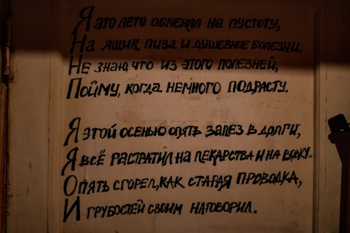 A wall features Russian text written in a bold, black script. It appears to be a poem or lyrical passage, and the writing is formatted in a visually striking manner with varying line lengths. The background is a muted beige color, giving a somber and subdued appearance. A shadow cast to the right covers part of the wall.
