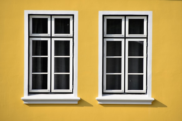 Two white-framed windows with multiple panes are set against a vibrant yellow wall. The windows have a symmetrical design and cast subtle shadows on the wall, suggesting bright daylight.