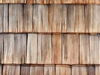 A close-up view of wooden shingles arranged in overlapping rows, displaying a natural weathered texture and varying shades of brown and gray. The wood grain is clearly visible, highlighting the rustic and aged appearance.
