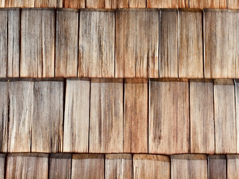A close-up view of wooden shingles arranged in overlapping rows, displaying a natural weathered texture and varying shades of brown and gray. The wood grain is clearly visible, highlighting the rustic and aged appearance.