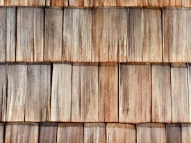 A close-up view of wooden shingles arranged in overlapping rows, displaying a natural weathered texture and varying shades of brown and gray. The wood grain is clearly visible, highlighting the rustic and aged appearance.