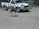 An ostrich stands on a gravel surface near a zebra-striped safari truck. The truck has a wooden bumper and displays an 'African Lion Safari' sign. In the background, there is a fence and some greenery.