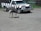 An ostrich stands on a gravel surface near a zebra-striped safari truck. The truck has a wooden bumper and displays an 'African Lion Safari' sign. In the background, there is a fence and some greenery.