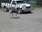 An ostrich stands on a gravel surface near a zebra-striped safari truck. The truck has a wooden bumper and displays an 'African Lion Safari' sign. In the background, there is a fence and some greenery.