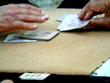 Two hands interacting with a deck of playing cards on a soft beige surface. One hand appears to be dealing a card while the other is placed above the cards as if to stop or collect them. The playing cards have intricate patterns, and colorful designs are visible on some cards.