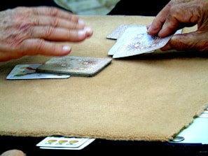 Two hands interacting with a deck of playing cards on a soft beige surface. One hand appears to be dealing a card while the other is placed above the cards as if to stop or collect them. The playing cards have intricate patterns, and colorful designs are visible on some cards.