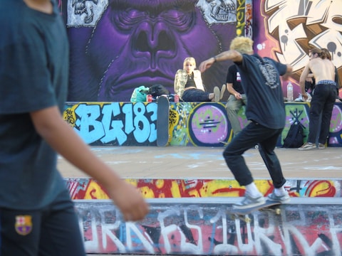 A vibrant urban scene featuring a skateboarder in motion on a graffiti-adorned skate park. Several individuals are gathered, relaxed and socializing, with a large mural depicting a stylized gorilla face in the background. The atmosphere conveys a youthful and energetic vibe.