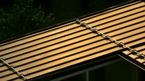 A metal roof with parallel lines reflecting a warm, golden light. The roof structure is visible with brackets or fasteners along the seams. In the background, there is a glimpse of green foliage, likely from nearby trees or plants.