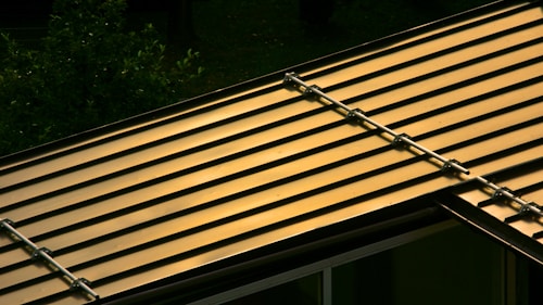 A metal roof with parallel lines reflecting a warm, golden light. The roof structure is visible with brackets or fasteners along the seams. In the background, there is a glimpse of green foliage, likely from nearby trees or plants.