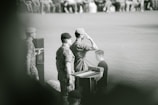 A group of military personnel are at a podium, with one person saluting. They are wearing camouflage uniforms and berets. The setting appears to be a formal ceremony or event, with an audience in the background.
