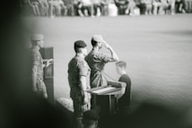 A group of military personnel are at a podium, with one person saluting. They are wearing camouflage uniforms and berets. The setting appears to be a formal ceremony or event, with an audience in the background.
