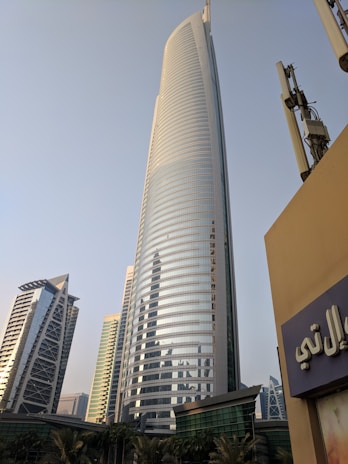 A tall, modern skyscraper with a sleek, curved design dominates the foreground, flanked by other high-rise buildings. In the bottom foreground, there are palm trees and a building with Arabic script on a sign. The sky is clear and slightly hazy.