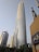 A tall, modern skyscraper with a sleek, curved design dominates the foreground, flanked by other high-rise buildings. In the bottom foreground, there are palm trees and a building with Arabic script on a sign. The sky is clear and slightly hazy.