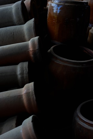 A collection of large, weathered clay pots and vases, some lying horizontally and others standing upright. The lighting is dim, creating deep shadows that enhance the textures and earthy tones of the pottery.