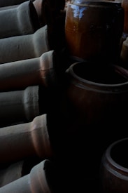 A collection of large, weathered clay pots and vases, some lying horizontally and others standing upright. The lighting is dim, creating deep shadows that enhance the textures and earthy tones of the pottery.