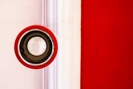 A circular speaker or light fixture with a red outer rim and a central white bulb is mounted against a wall. The background is divided into a bright white section on the left and a vibrant red section on the right.