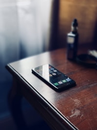A smartphone with a dark screen is lying face up on a wooden table, next to a vape pen. The sunlight from a window casts a soft glow, creating reflections on the phone's screen. The surroundings have a warm, rustic ambiance.