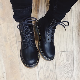 Black leather boots with yellow stitching are worn over dark pants, standing on a light wooden floor.