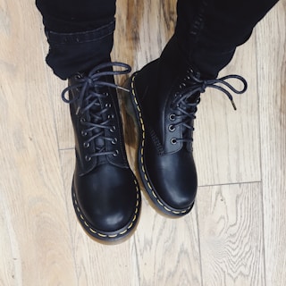 Black leather boots with yellow stitching are worn over dark pants, standing on a light wooden floor.
