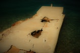 A broken and cracked concrete pier juts into a body of water. The pier has large holes and visible decay, with sections of rebar and rubble exposed. The water surrounding the pier is clear and greenish-blue, hinting at a shallow depth. The overall structure appears weathered and worn by the elements.