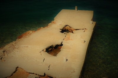 A broken and cracked concrete pier juts into a body of water. The pier has large holes and visible decay, with sections of rebar and rubble exposed. The water surrounding the pier is clear and greenish-blue, hinting at a shallow depth. The overall structure appears weathered and worn by the elements.