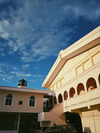 The image displays a mosque with a light beige facade and multiple arched windows. There is a smaller building with a dome on top, which contrasts against the vibrant blue sky filled with scattered clouds. The sunlight casts shadows, accentuating the architectural details.