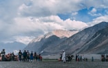 A group of people and camels are gathered on a sandy expanse with a backdrop of majestic mountain ranges under a partly cloudy sky. Some individuals appear to be tourists, possibly engaging in a camel ride, while others stand nearby observing the scene. The landscape conveys a sense of openness and natural beauty.