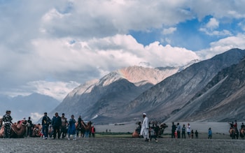 A group of people and camels are gathered on a sandy expanse with a backdrop of majestic mountain ranges under a partly cloudy sky. Some individuals appear to be tourists, possibly engaging in a camel ride, while others stand nearby observing the scene. The landscape conveys a sense of openness and natural beauty.