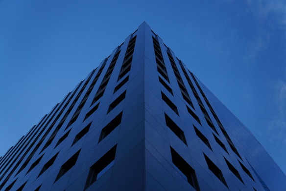 A modern, angular skyscraper photographed from a low angle, highlighting its geometric design and vertical lines. The structure appears to be made of reflective glass, enhancing its sleek and contemporary appearance. The surrounding sky is a clear, deep blue, providing a striking contrast to the dark hues of the building.