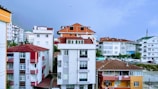 A cityscape featuring multiple apartment buildings with varying architectural styles. The central building has a modern design with a white facade and large windows, while others have red and white elements or unfinished brickwork. Cars are parked along the street, and a minaret can be seen in the distance, suggesting the presence of a mosque.