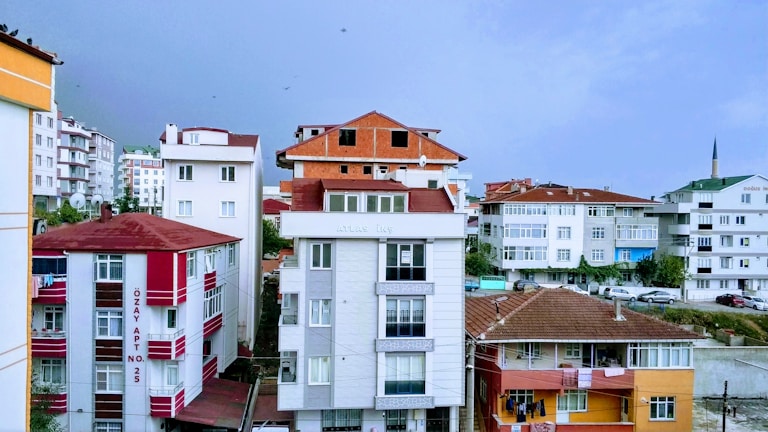 A cityscape featuring multiple apartment buildings with varying architectural styles. The central building has a modern design with a white facade and large windows, while others have red and white elements or unfinished brickwork. Cars are parked along the street, and a minaret can be seen in the distance, suggesting the presence of a mosque.
