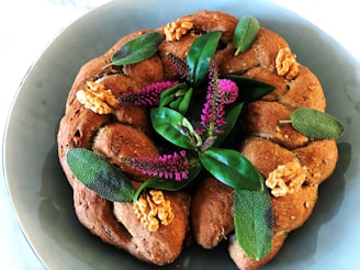 A braided round loaf of bread adorned with leafy greens, purple flowers, and walnuts. The bread is placed on a gray plate, showcasing a decorative and rustic presentation.