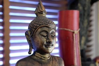 A golden statue of the Buddha with a serene expression is positioned against a background with horizontal wooden blinds. The statue features intricate detailing on the head and facial features, while a red cylindrical object tied with twine is partially visible to the side.