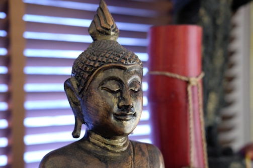A golden statue of the Buddha with a serene expression is positioned against a background with horizontal wooden blinds. The statue features intricate detailing on the head and facial features, while a red cylindrical object tied with twine is partially visible to the side.