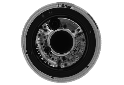 A detailed x-ray image of a circular object revealing intricate internal components and circuitry. The symmetrical arrangement shows various circular elements and patterns, likely denoting mechanical or electronic parts.