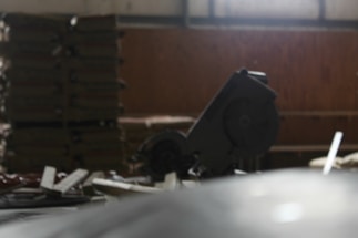 A dimly lit workshop or construction area featuring a circular saw in the foreground. Stacks of materials, possibly wooden boards, can be seen in the background along with assorted tools and debris scattered across the surface.