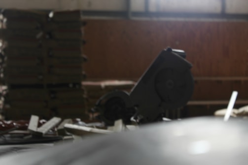 A dimly lit workshop or construction area featuring a circular saw in the foreground. Stacks of materials, possibly wooden boards, can be seen in the background along with assorted tools and debris scattered across the surface.