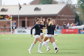 Three women in black athletic shirts and white shorts are on a grassy field, engaged in some activity that appears to be related to sports practice or warm-up. They are in an open area with a building with large windows in the background. There are also a few people visible near the building. The field is marked for a sporting event, and a few pieces of sports equipment can be seen, including a soccer ball and some training cones.