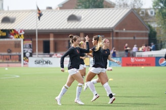 Three women in black athletic shirts and white shorts are on a grassy field, engaged in some activity that appears to be related to sports practice or warm-up. They are in an open area with a building with large windows in the background. There are also a few people visible near the building. The field is marked for a sporting event, and a few pieces of sports equipment can be seen, including a soccer ball and some training cones.