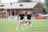 Three women in black athletic shirts and white shorts are on a grassy field, engaged in some activity that appears to be related to sports practice or warm-up. They are in an open area with a building with large windows in the background. There are also a few people visible near the building. The field is marked for a sporting event, and a few pieces of sports equipment can be seen, including a soccer ball and some training cones.
