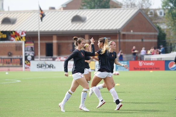 Three women in black athletic shirts and white shorts are on a grassy field, engaged in some activity that appears to be related to sports practice or warm-up. They are in an open area with a building with large windows in the background. There are also a few people visible near the building. The field is marked for a sporting event, and a few pieces of sports equipment can be seen, including a soccer ball and some training cones.