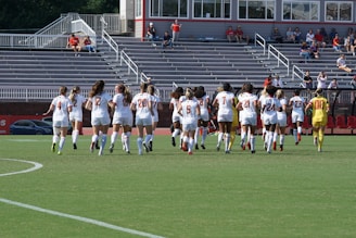 A group of female soccer players in white jerseys are jogging on a field towards empty bleachers in a stadium. Each player wears a number on her back, and the goalie's outfit is distinctively yellow. Some spectators are visible in the stands, with a few people watching the event.