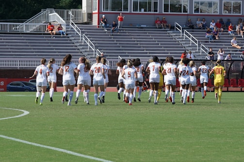 A group of female soccer players in white jerseys are jogging on a field towards empty bleachers in a stadium. Each player wears a number on her back, and the goalie's outfit is distinctively yellow. Some spectators are visible in the stands, with a few people watching the event.